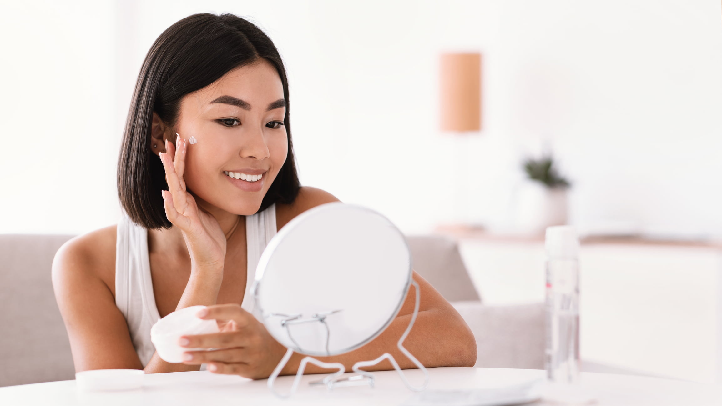 A pretty Asian lady is looking at the mirror and applying a skincare cream